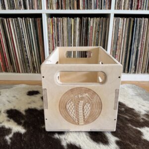 Baltic birch wood 7″ vinyl crate showing laser‑engraved cobra logo on one side, placed on a cowhide rug with vinyl records in the background.